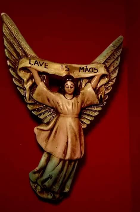 Anjo de gesso pendurado na parede vermelha segurando uma faixa onde se lê “Lave as mãos”.